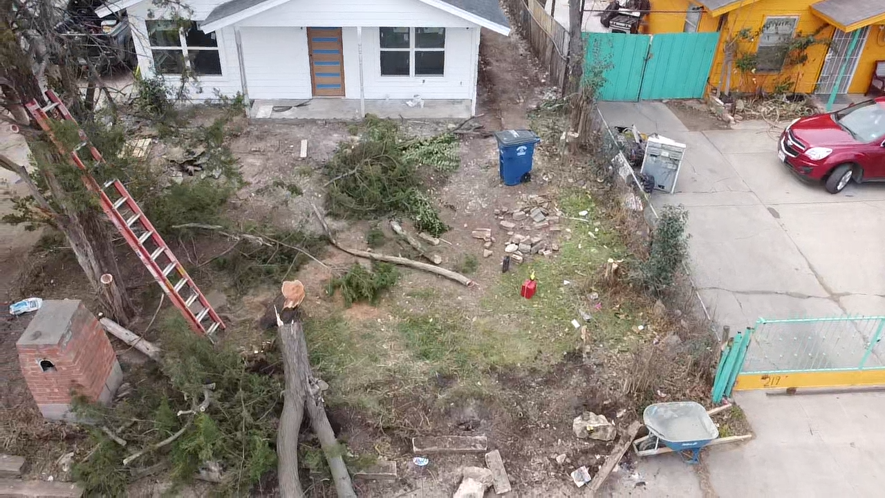 Straight-on view of the cluttered front lot showing cut stumps, a leaning ladder, broken concrete, and a wheelbarrow before the yard rebuild